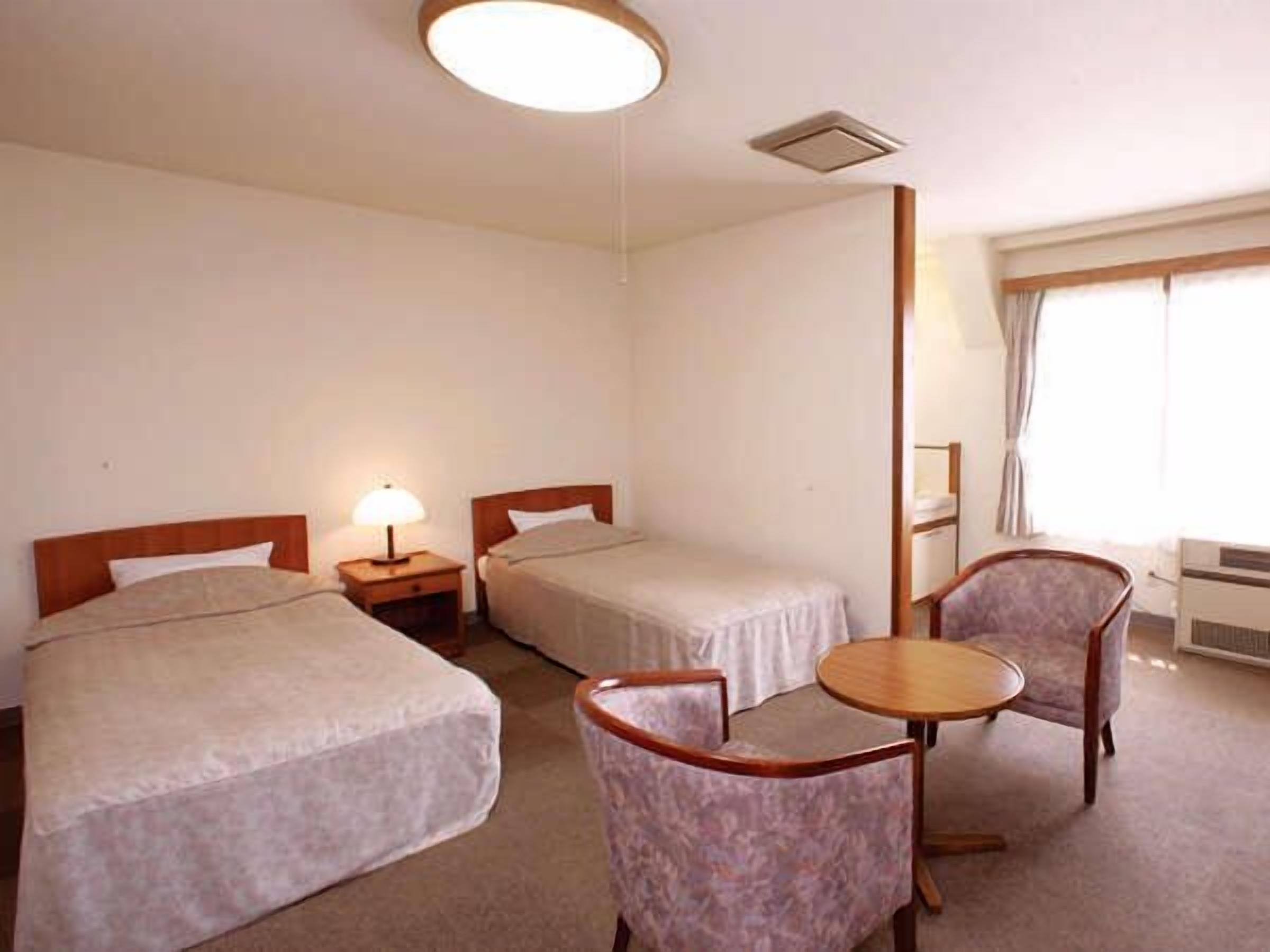 A hotel room with two beds, a small table, a chair, and a window with curtains.