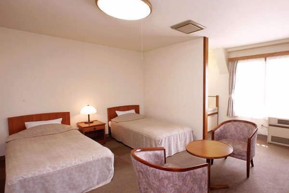 Ein Hotelzimmer mit zwei Betten, einem kleinen Tisch, einem Stuhl und einem Fenster mit Vorhängen.