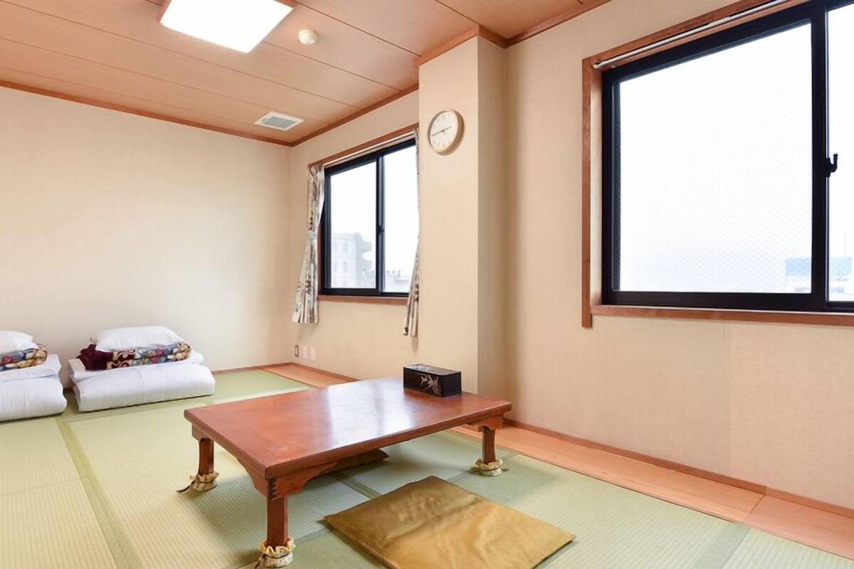 Ein Zimmer mit Tatami-Matten, einem Holztisch, zwei Kissen, einer Uhr und einem Fenster.