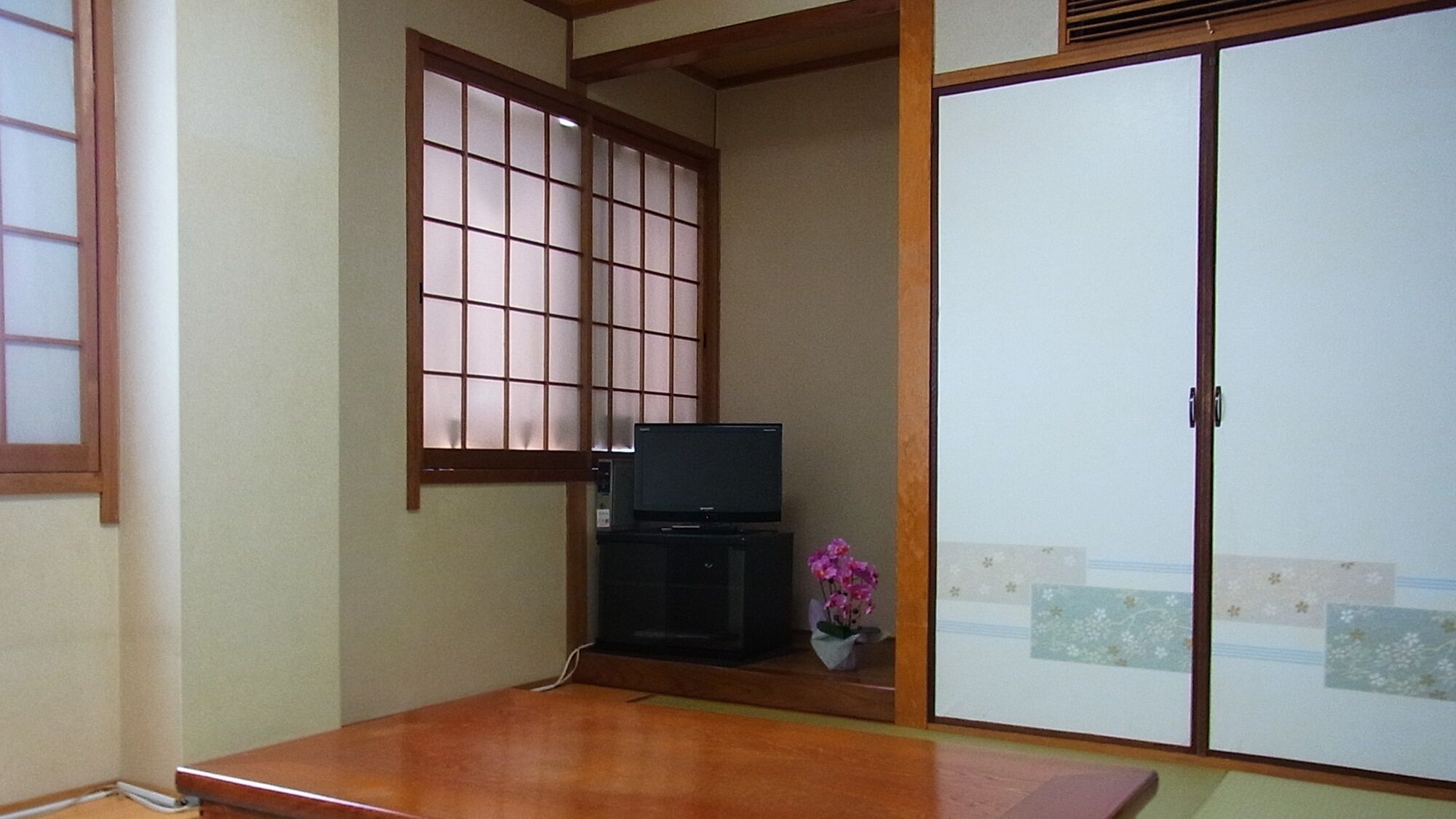Une pièce de style japonais traditionnel, avec un sol recouvert de tatamis, une porte coulissante, un téléviseur posé sur un meuble bas et un petit arrangement floral.
