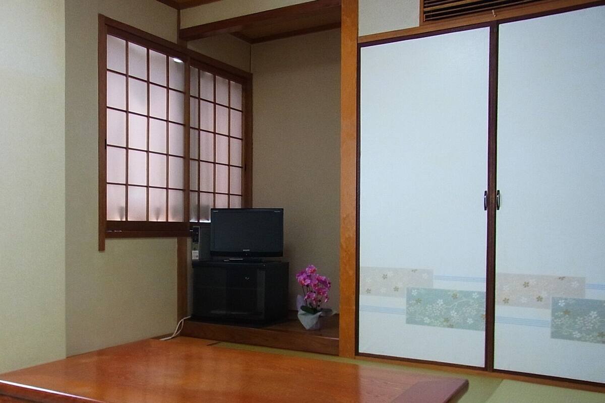 Ein traditioneller japanischer Raum mit Tatami-Bodenbelag, einer Schiebetür, einem Fernseher auf einem niedrigen Schrank und einer kleinen Blumenkomposition.