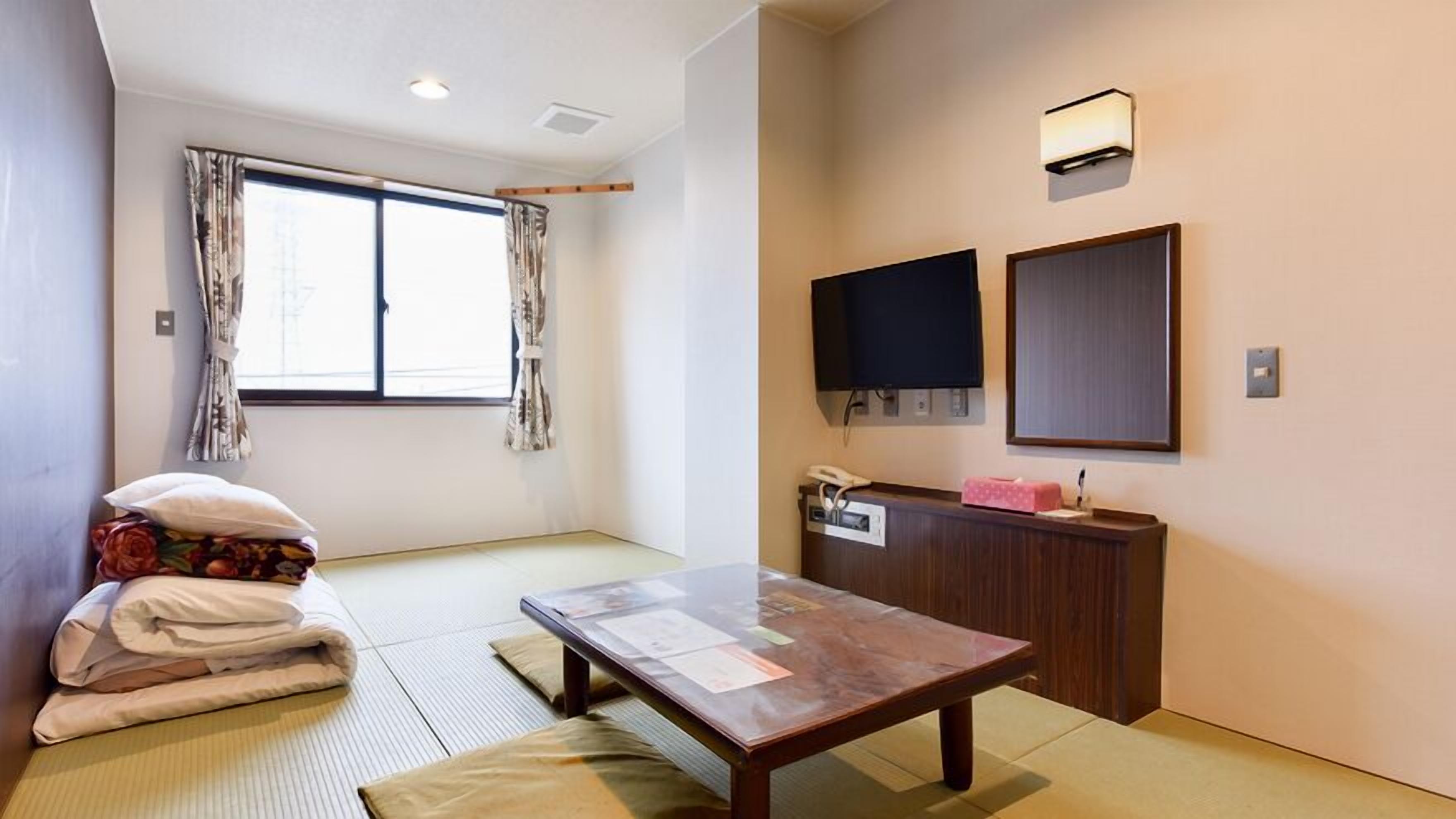 Een kamer met tatami vloerbedekking, een lage tafel, een bed met kussens, een bureau met telefoon en twee wandtelevisies.