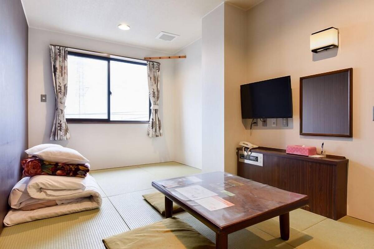 Ein Zimmer mit Tatami-Boden, einem niedrigen Tisch, einem Bett mit Kissen, einem Schreibtisch mit Telefon und zwei Wand-Fernsehern.