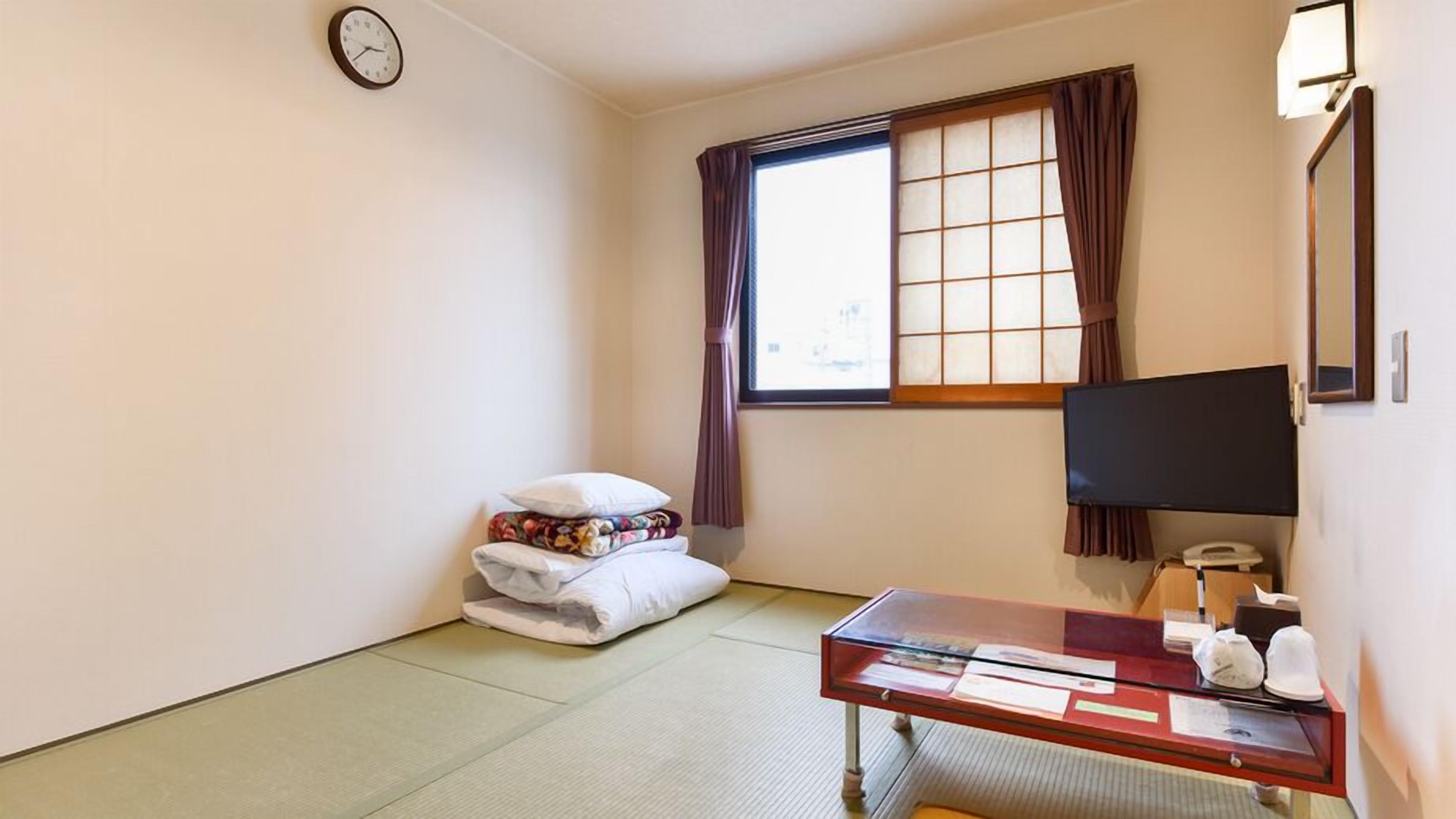 Een hotelkamer met tatami vloeren, een bed met kussens, een bureau met een computer, een televisie en een raam met gordijnen.