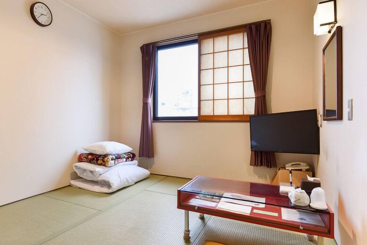 Ein Hotelzimmer mit Tatami-Boden, einem Bett mit Kissen, einem Schreibtisch mit Computer, einem Fernseher und einem Fenster mit Vorhängen.