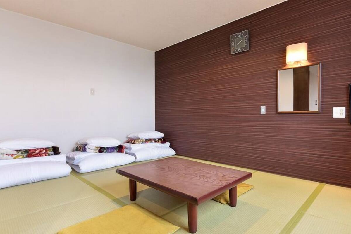 Ein Zimmer mit Tatami-Boden, einem niedrigen Holztisch und einem an der Wand befestigten Fernseher.