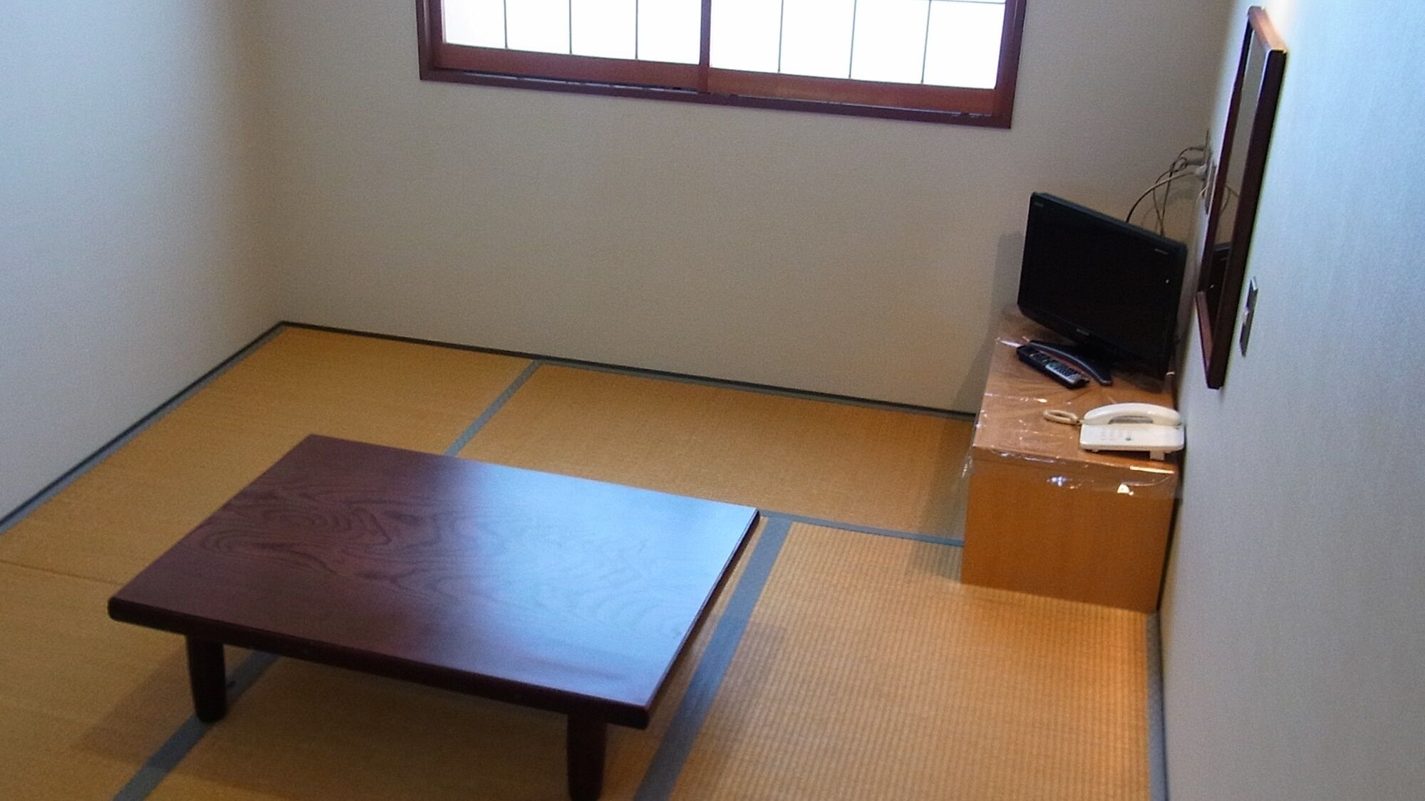 Une pièce avec un sol recouvert de tatamis, une petite table en bois, un téléviseur posé sur un meuble et un téléphone.