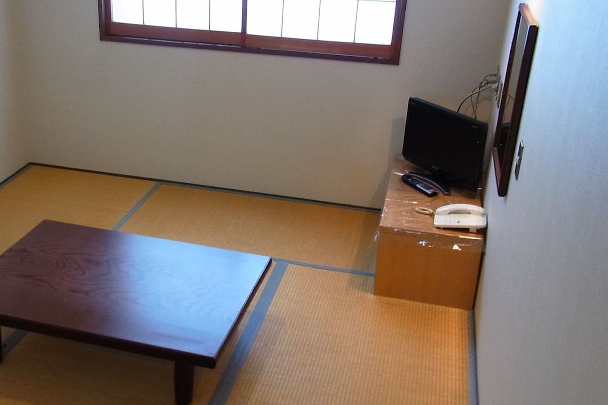 Una habitación con suelo de tatami, una mesa de madera baja, un televisor sobre un mueble y un teléfono.