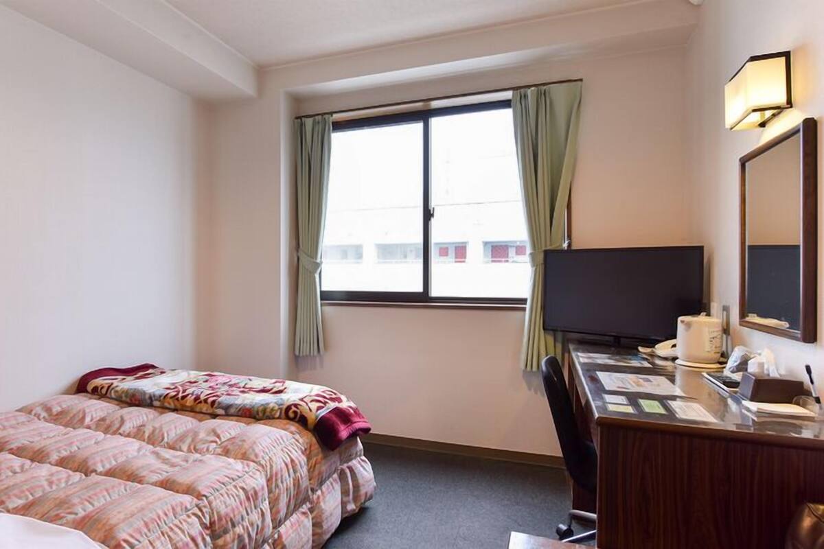 Ein Hotelzimmer mit Bett, Schreibtisch mit Computer, Fernseher und einem Fenster mit Vorhängen.