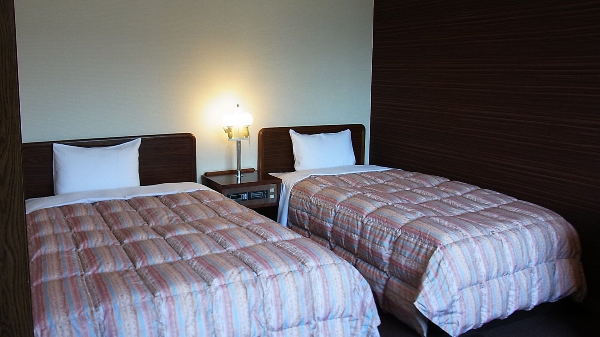 Deux lits avec des draps à rayures, une table de chevet avec une lampe et une tête de lit en bois.