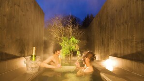 Una pareja en una bañera exterior climatizada, con una botella de champán y copas, rodeada de paredes de madera y un árbol.