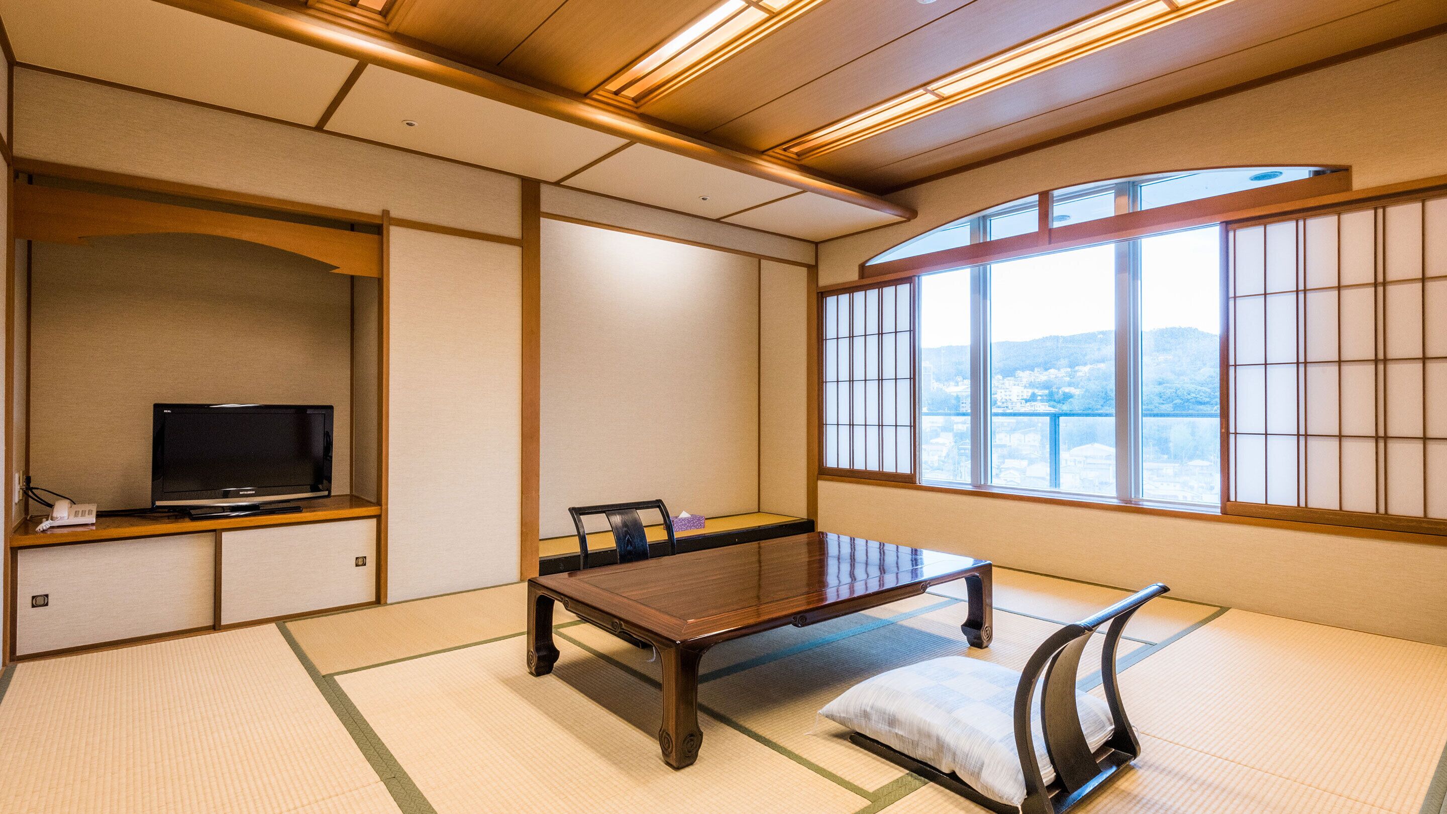 A traditional Japanese room with tatami flooring, a low table, a television on a shelf, and a view through large windows.