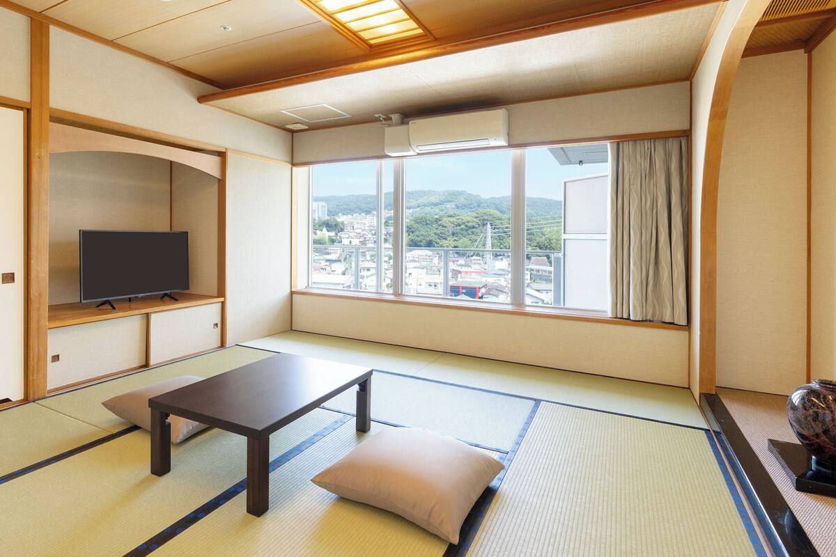Um quarto japonês tradicional com tatames, uma televisão em uma prateleira baixa, uma mesa com almofadas e uma grande janela com vista para a cidade.