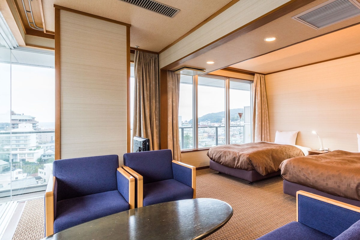 Quarto de hotel com duas camas, área de estar com mesa e grandes janelas com vista para a cidade.