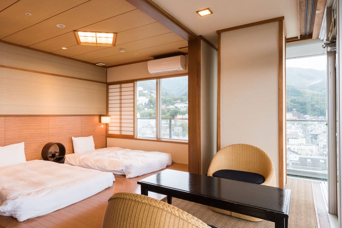 Quarto de hotel com duas camas, uma mesa pequena, uma cadeira e vista da cidade por grandes janelas.