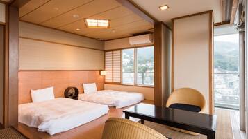 Habitación de hotel con dos camas, una mesita, una silla y vistas de la ciudad a través de amplias ventanas.