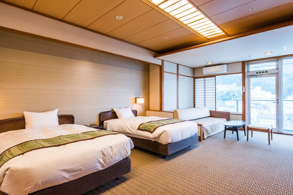 Quarto de hotel com duas camas, uma mesinha e vista para a cidade através de grandes janelas.