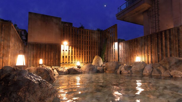 Un área de piscina al aire libre tranquila con estructuras de madera y rocas de piedra, iluminada con una luz cálida.