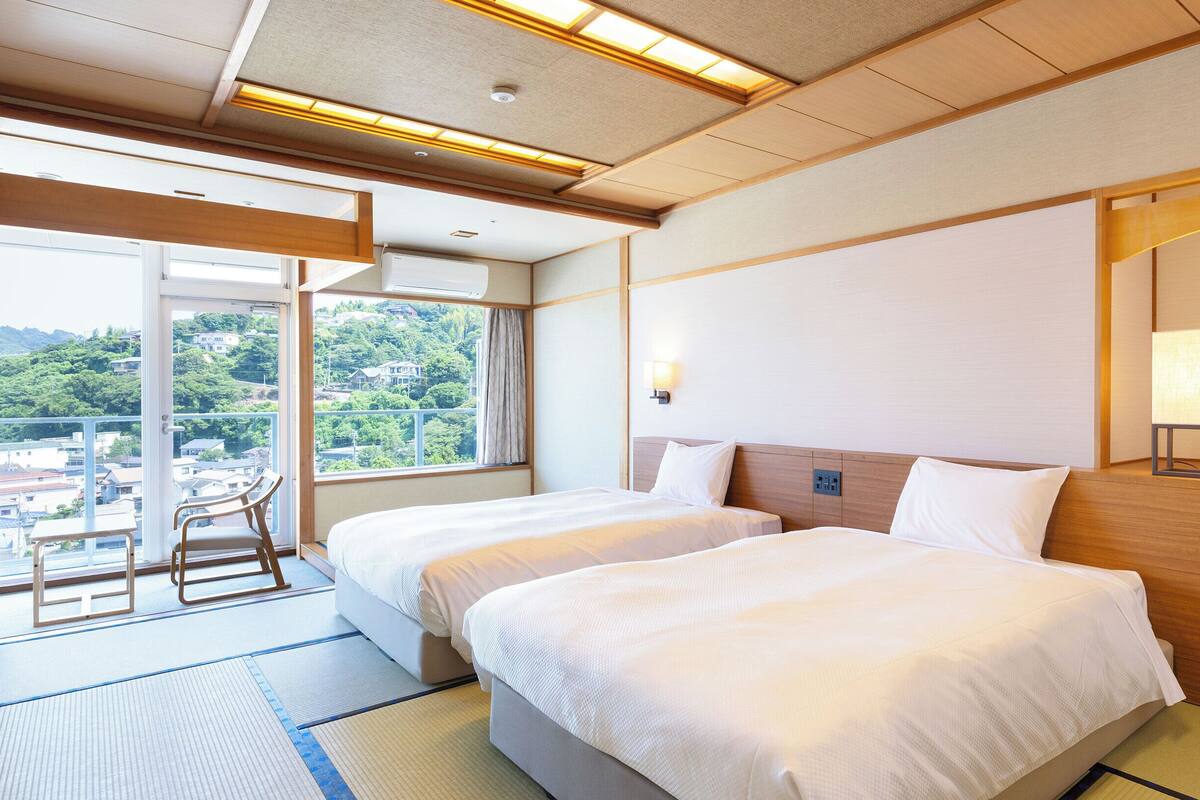 Quarto de hotel com duas camas, vista para a cidade, varanda com uma cadeira e um candeeiro de estilo tradicional japonês.