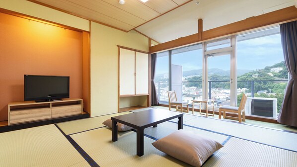 Una habitación de estilo japonés tradicional con suelo de tatami, televisor, mesa y vistas a la ciudad a través de amplias ventanas.