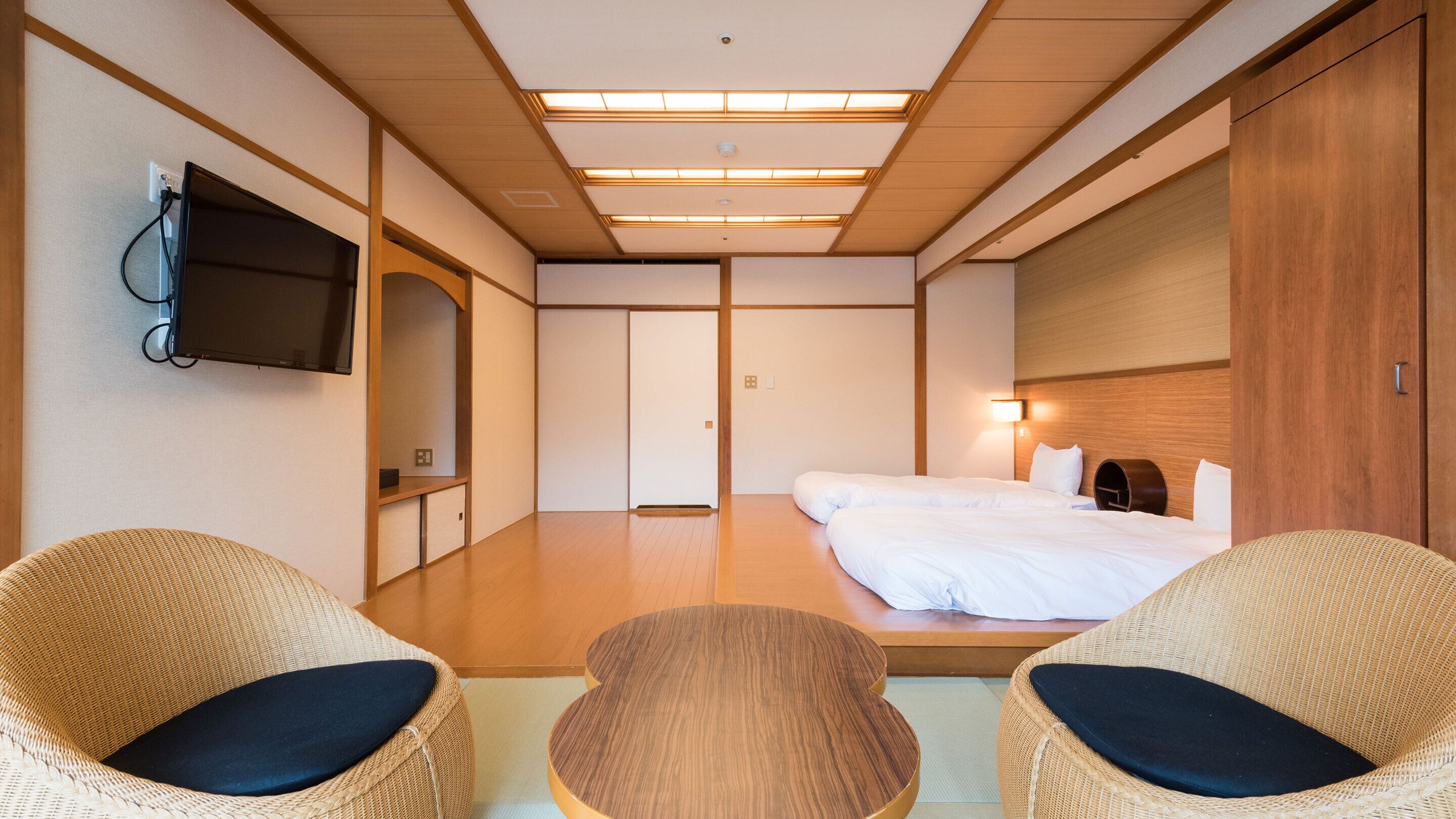 A hotel room with a bed, two wicker chairs, a television, and a wooden wardrobe.