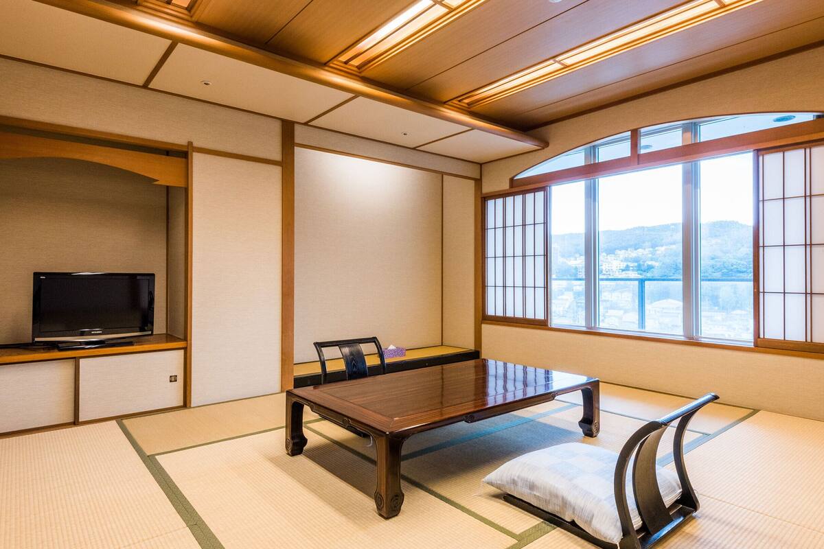 Um quarto japonês tradicional com tatames, uma mesa baixa, uma televisão numa prateleira e vista através de grandes janelas.