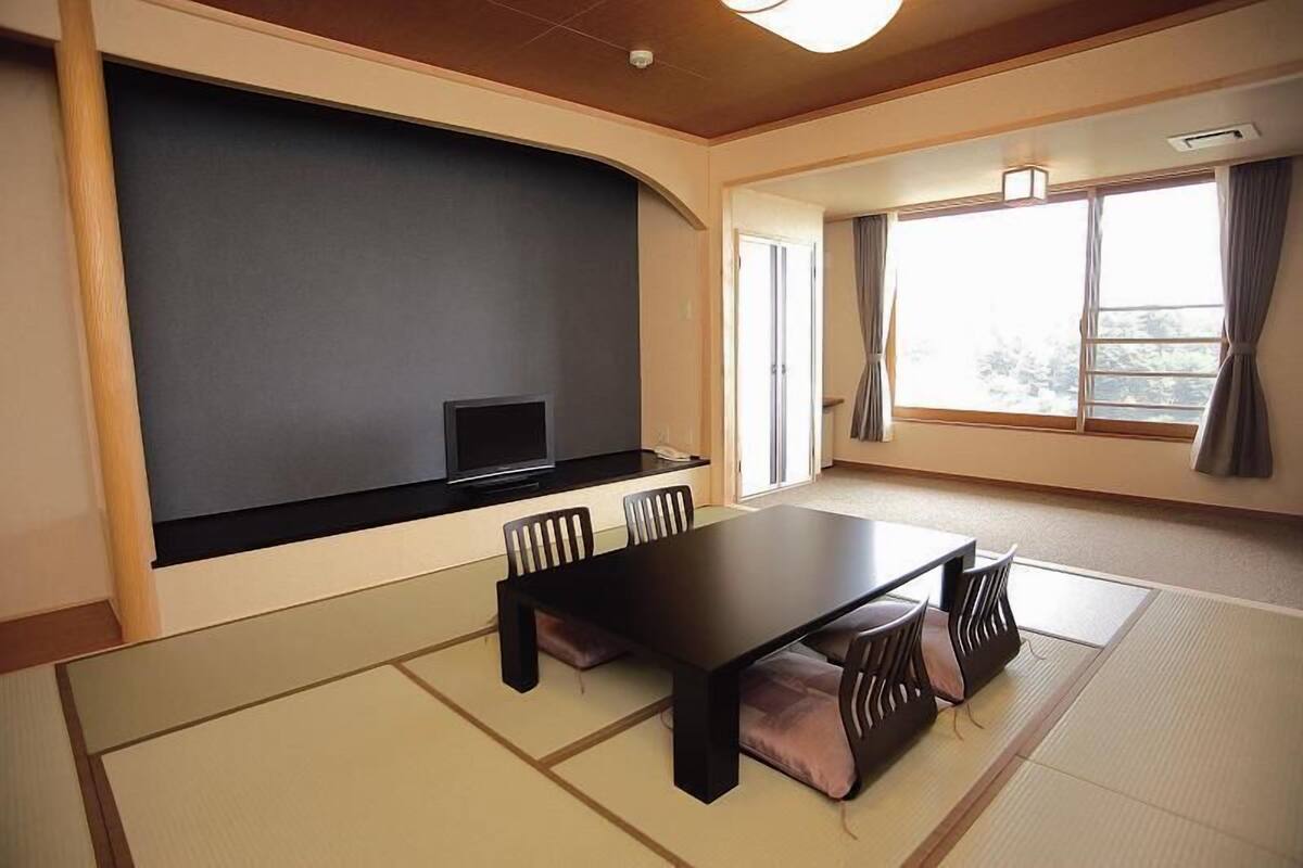 A room with tatami flooring, a dark wooden table, and chairs, a television on a low wall, and a large window with curtains.