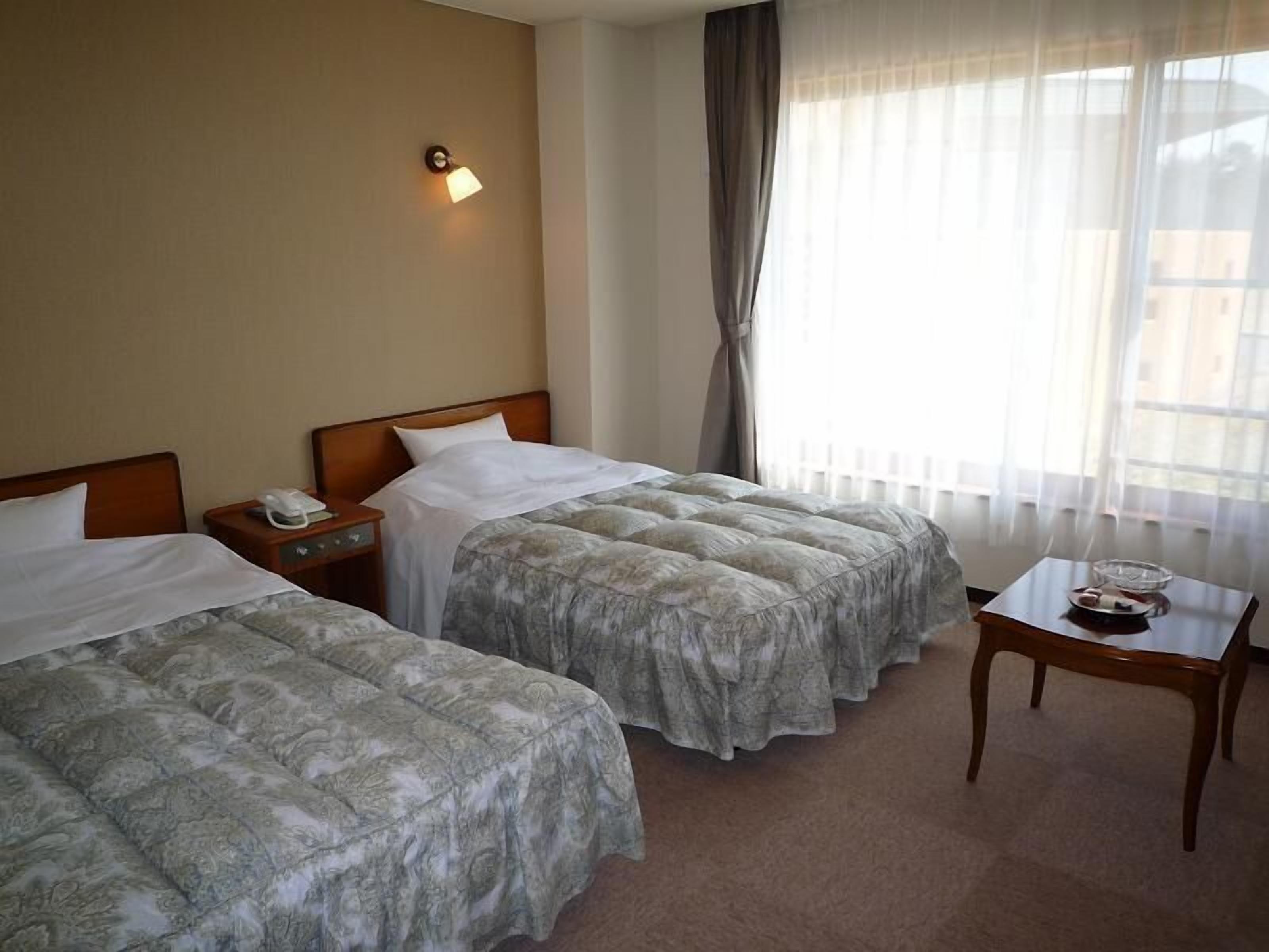 Habitación de hotel con dos camas, una mesita de noche con teléfono, una pequeña mesa con bandeja y una ventana con cortinas.