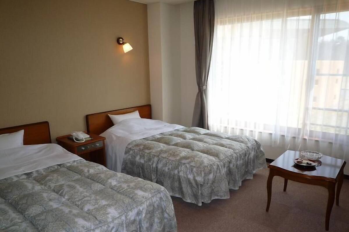 A hotel room with two beds, a nightstand with a phone, a small table with a tray, and a window with curtains.