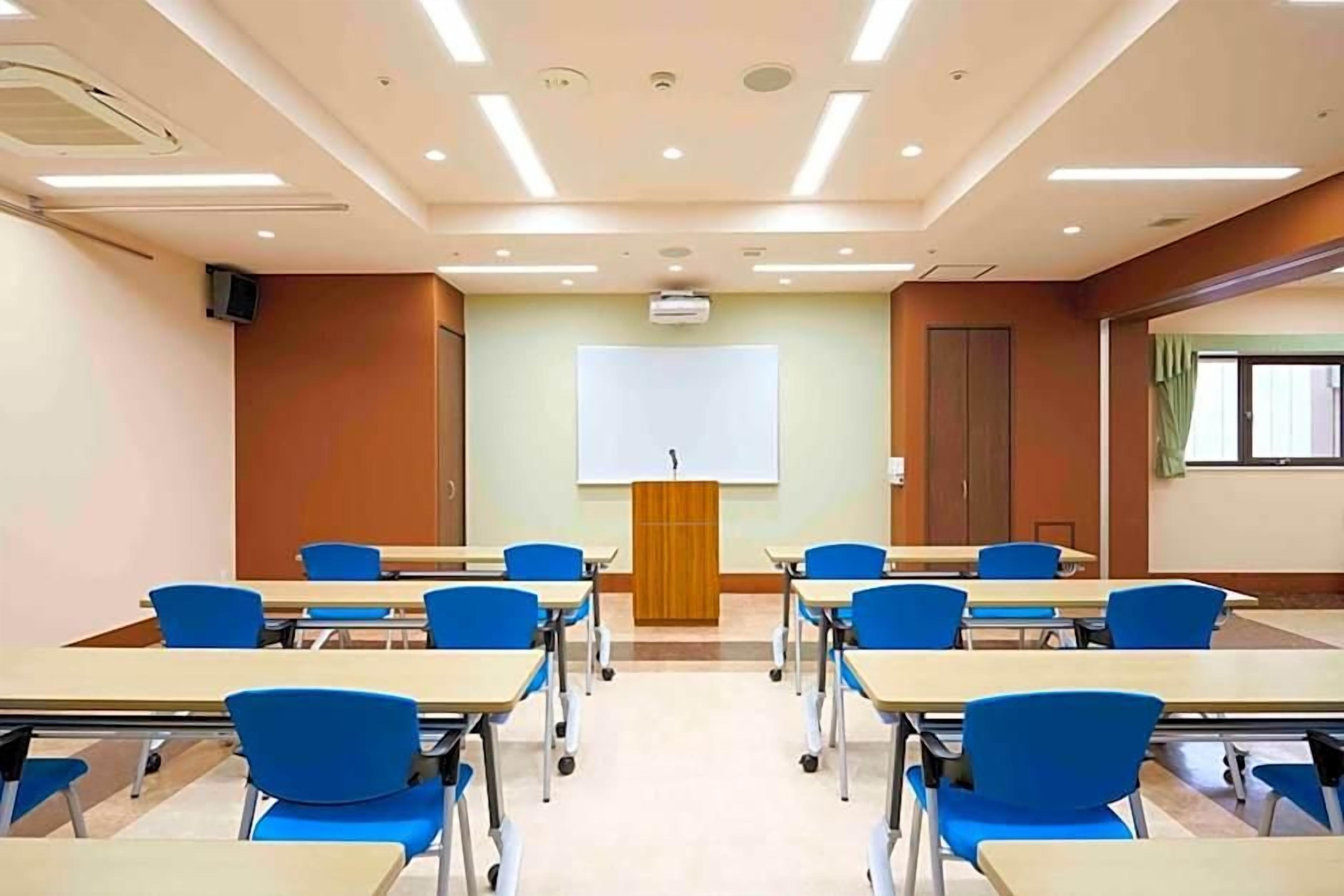 A classroom with desks and chairs arranged in rows, a projector screen at the front, and a door on the right.