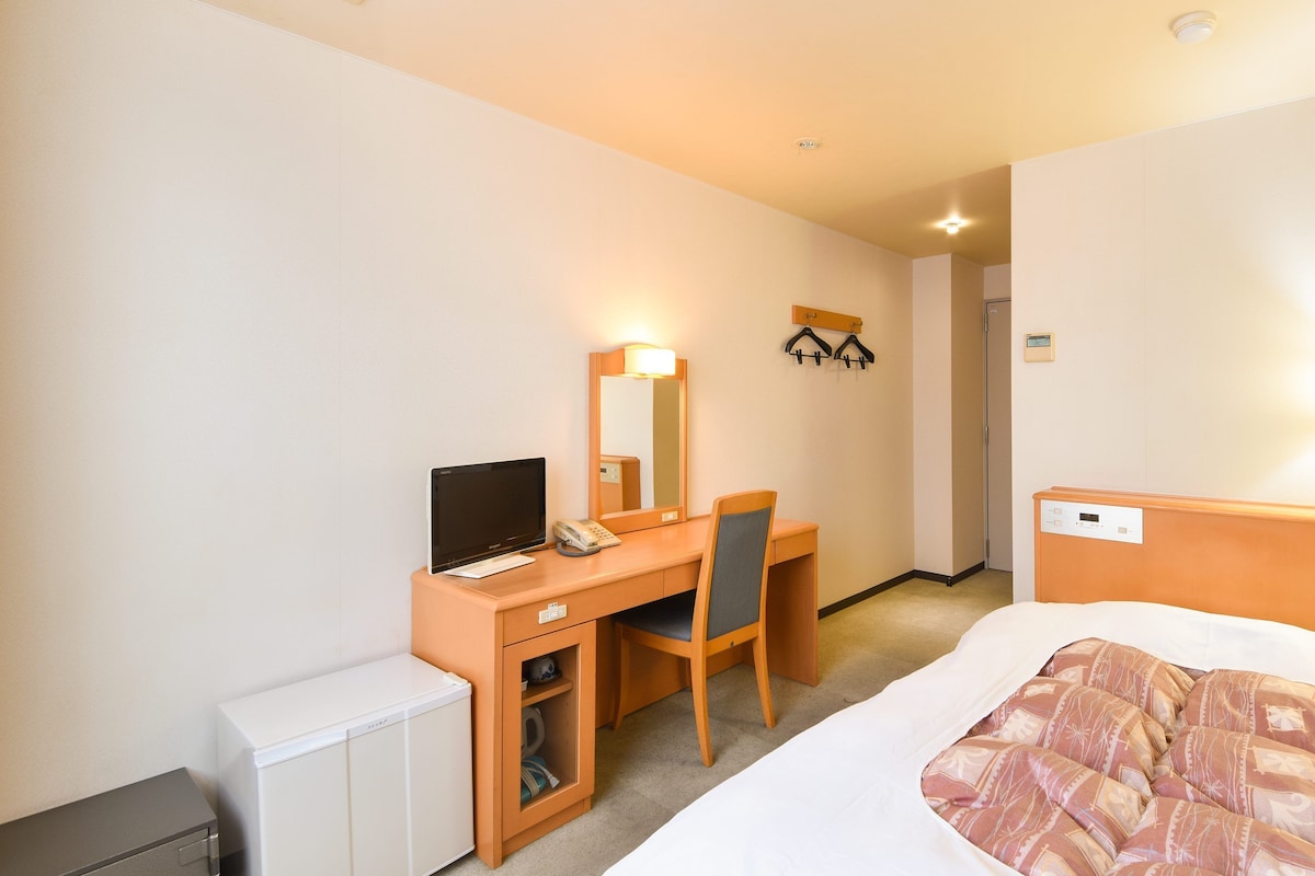 Una habitación de hotel con cama, escritorio con computadora, silla, refrigerador pequeño y mesita de noche.