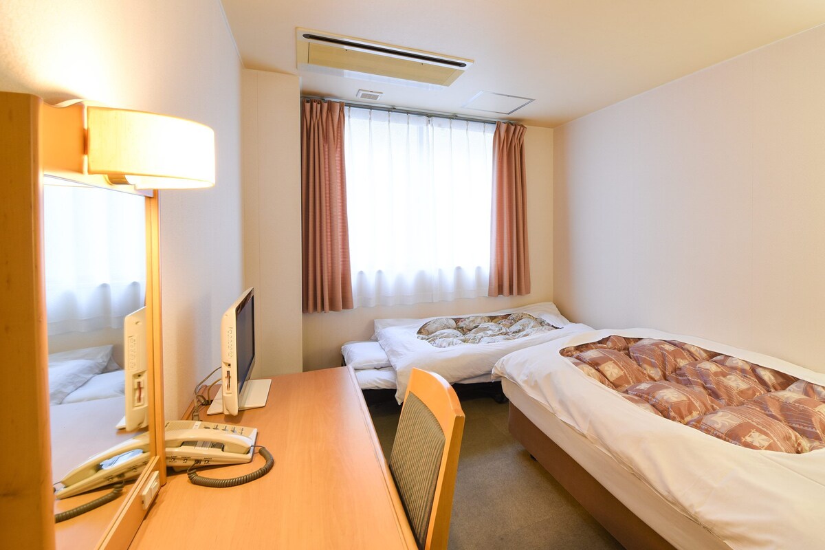 Habitación de hotel con escritorio, cama, silla, equipo de aire acondicionado y ventana con cortinas.