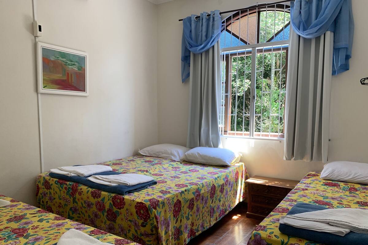 Ein Zimmer mit zwei Betten, jeweils mit Blumenmuster-Bettwäsche, einem Fenster mit Vorhängen, einem an der Wand befestigten Lichtschalter und einem gerahmten Bild.