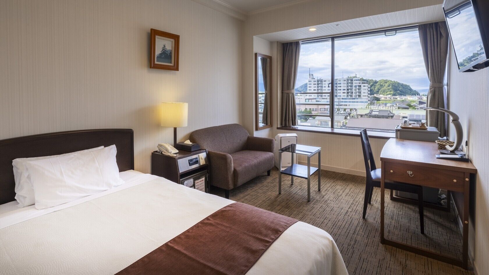 A hotel room with a bed, a desk, a chair, a lamp, a telephone, and a window with a city view.