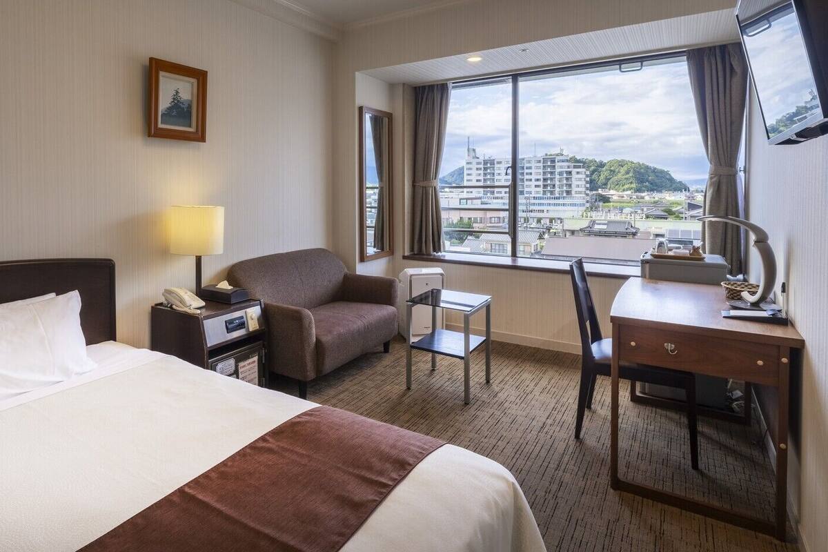 A hotel room with a bed, a desk, a chair, a lamp, a telephone, and a window with a city view.