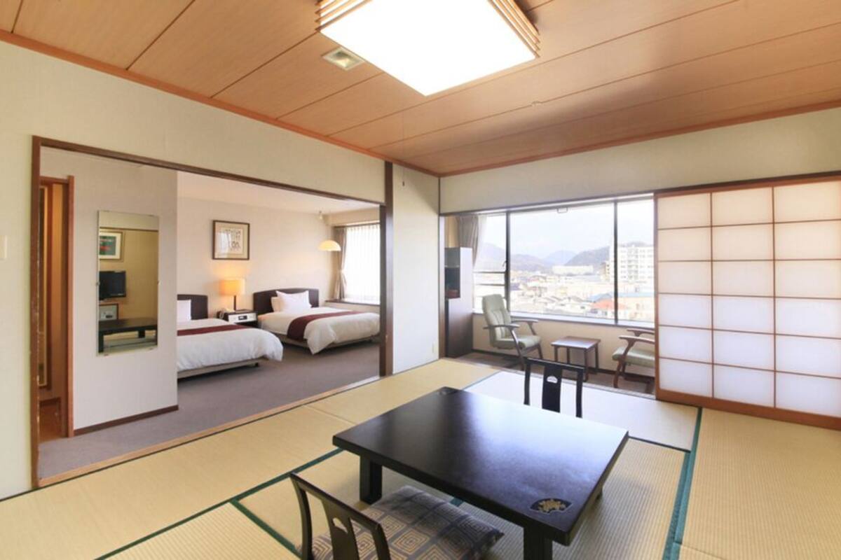 A traditional Japanese-style room with tatami flooring, a low table, a bed, and a sliding door leading to a balcony with a city view.