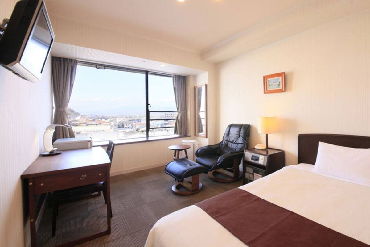 A hotel room with a bed, desk, chair, TV, and a window with a city view.