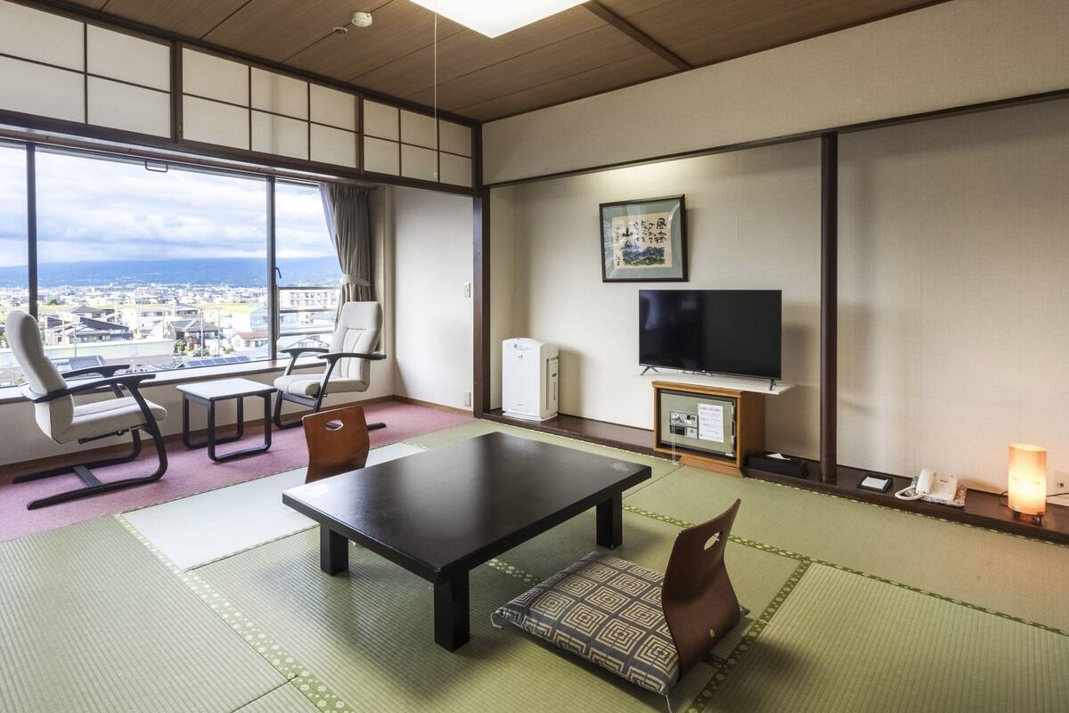 A room with tatami flooring, a television, a desk, chairs, and a view of a cityscape through large windows.