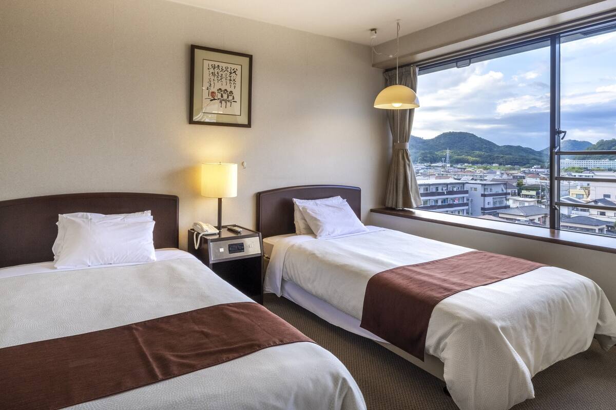 A hotel room with two beds, a nightstand with a lamp, a framed picture on the wall, and a window with a city view.
