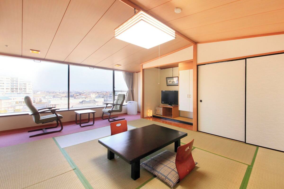 A modern hotel room with tatami mats, a large window with a city view, a flat-screen TV, and a sliding door.
