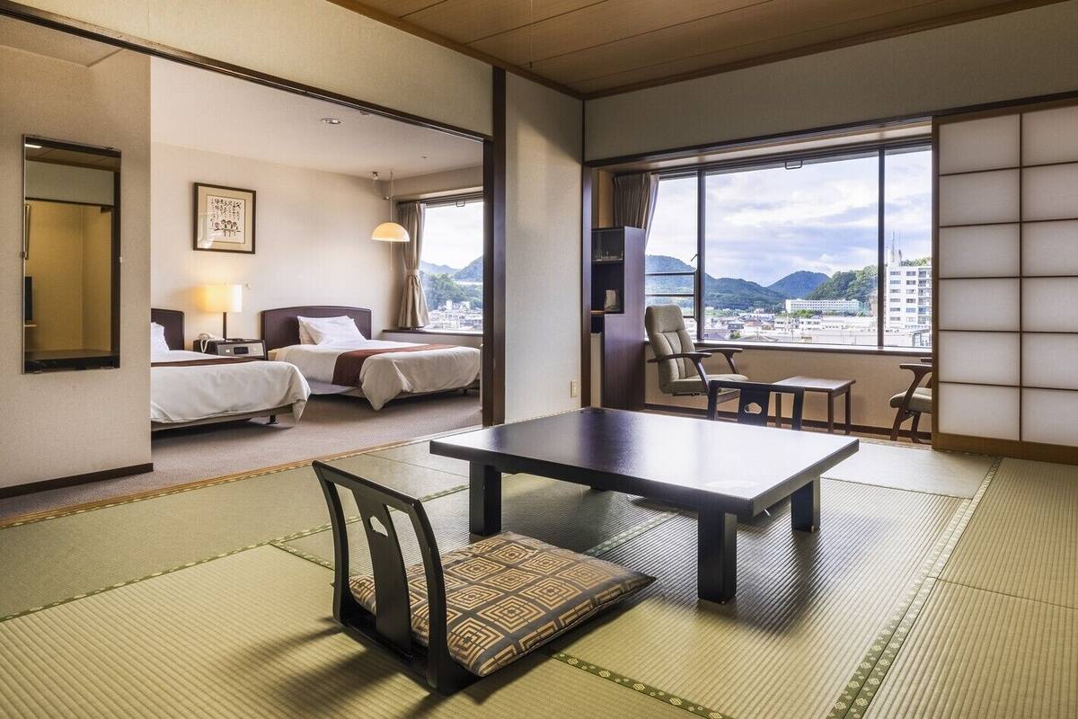 A traditional Japanese-style room with tatami flooring, a low table with chairs, a bed with white bedding, and a view of mountains through large windows.