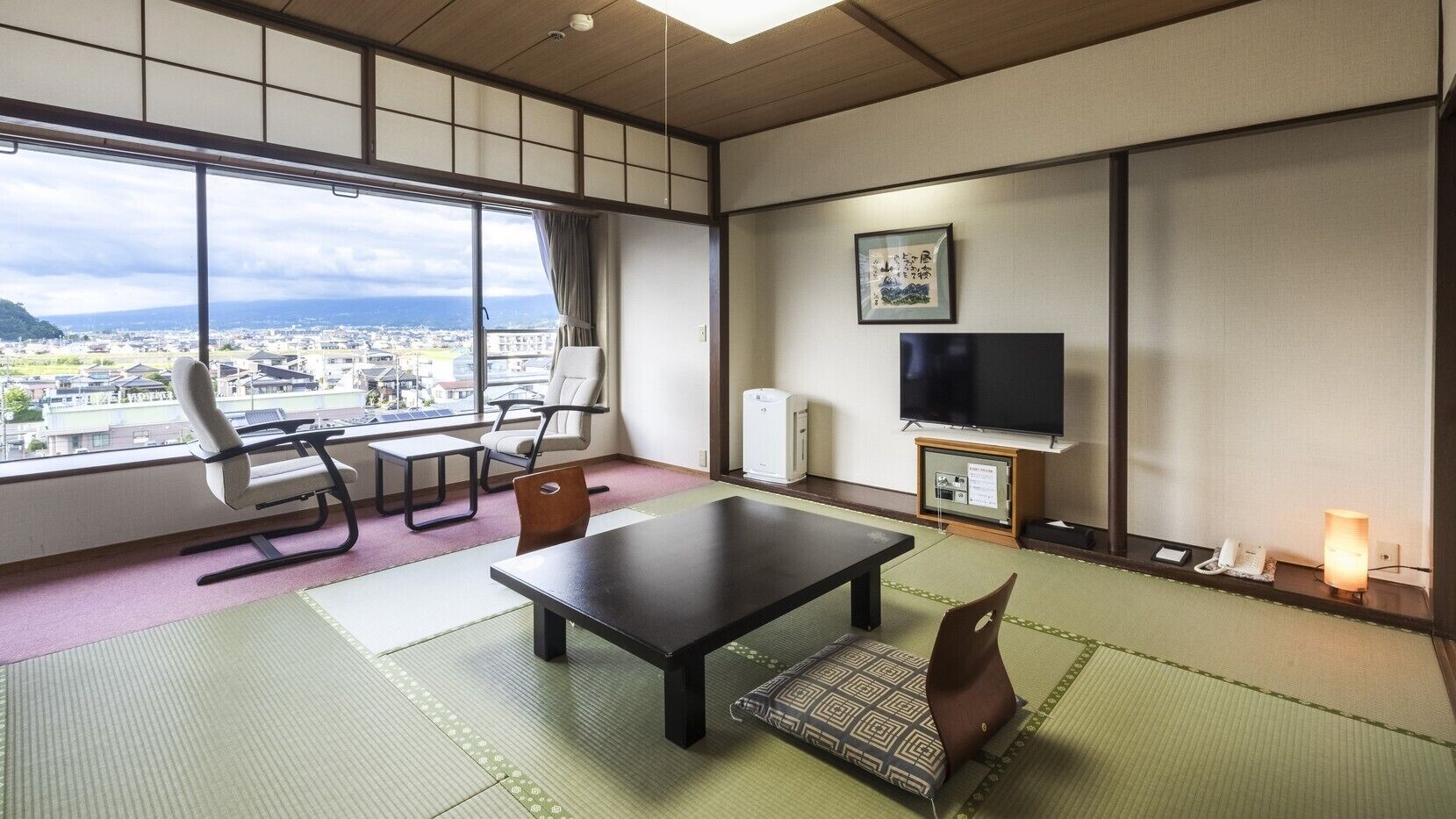 A room with tatami flooring, a table, chairs, a TV, a heater, and a view of a cityscape.