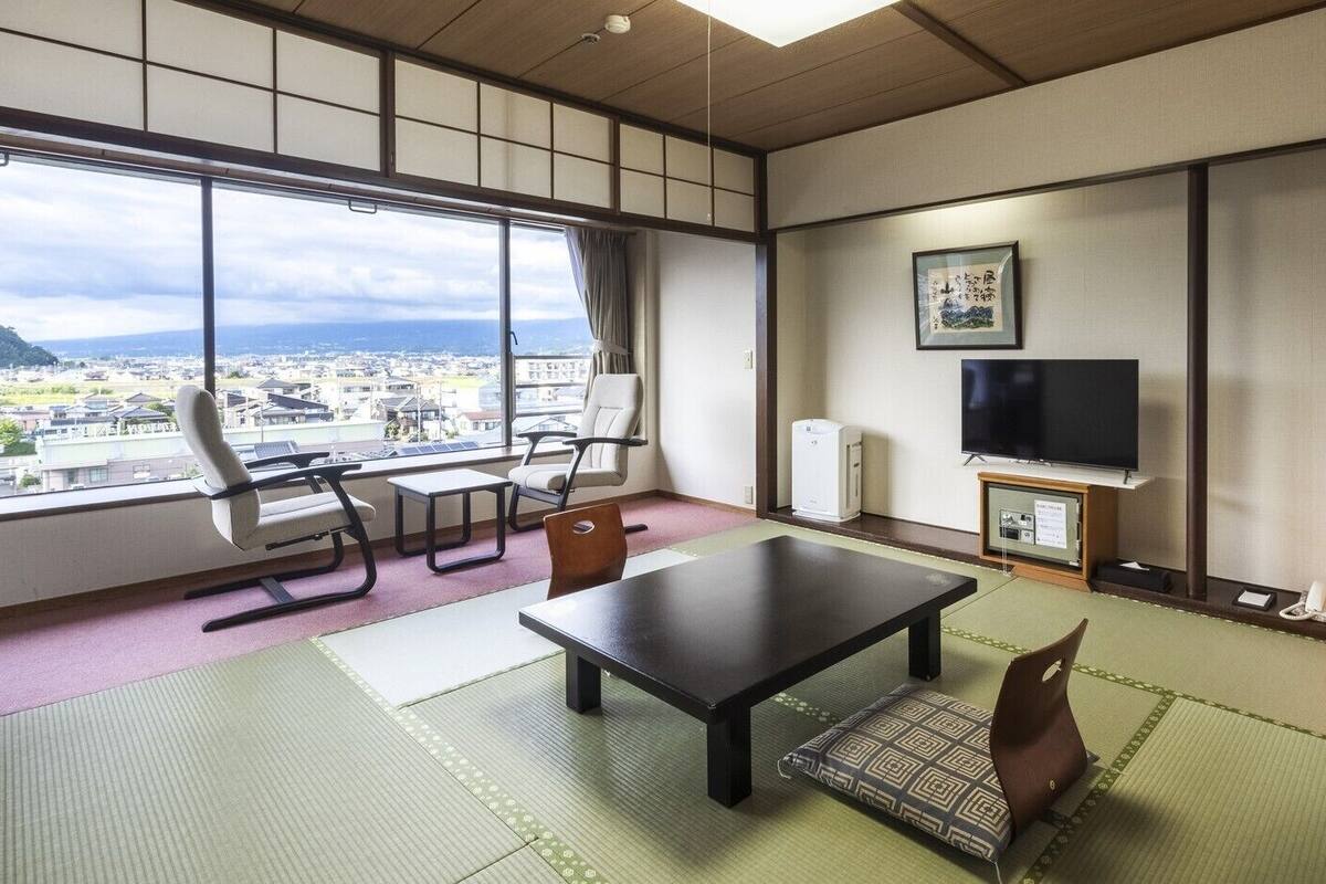 A room with tatami flooring, a table, chairs, a TV, a heater, and a view of a cityscape.