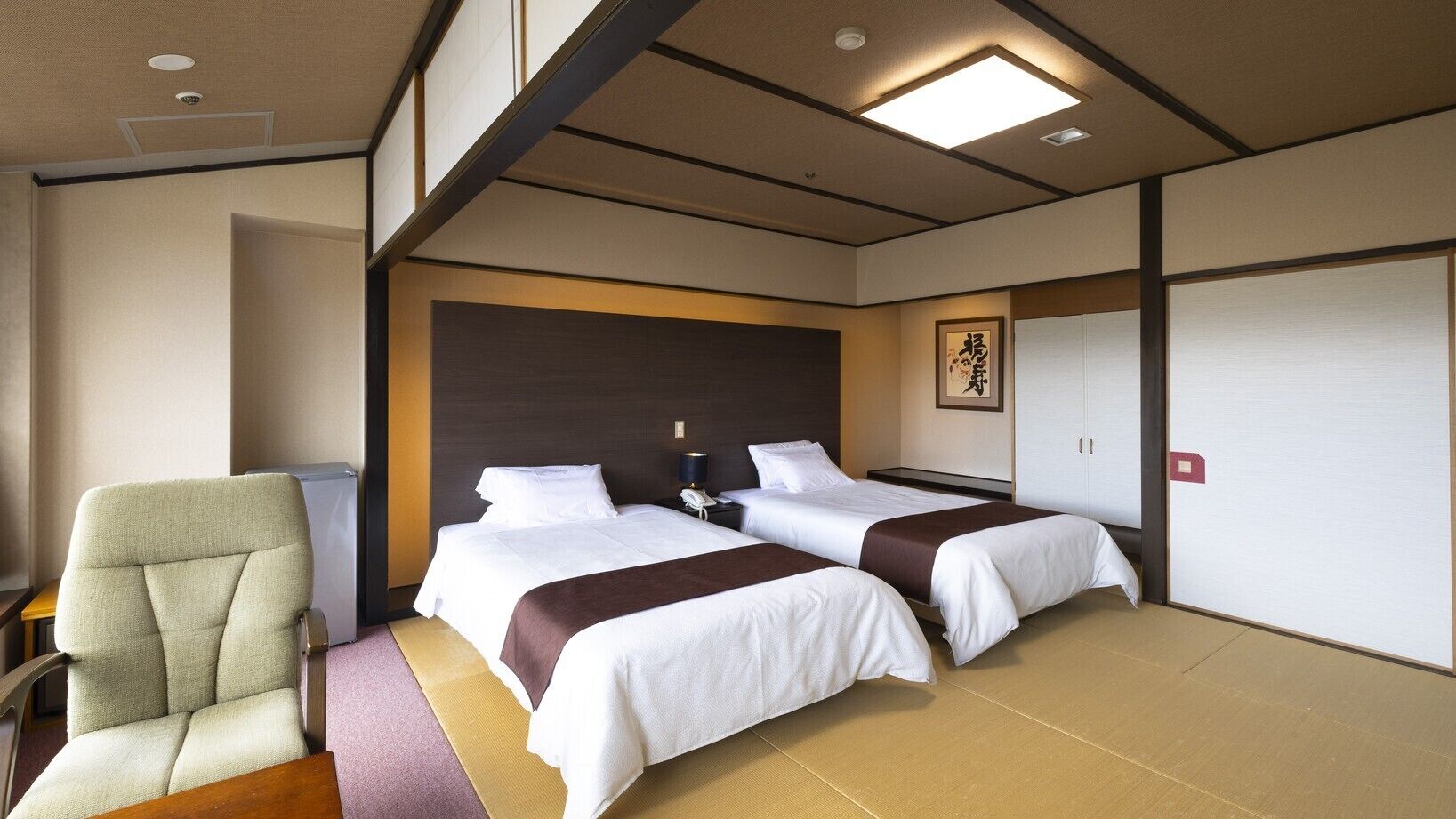 A hotel room with two beds, a chair, a desk, and a wall-mounted phone.