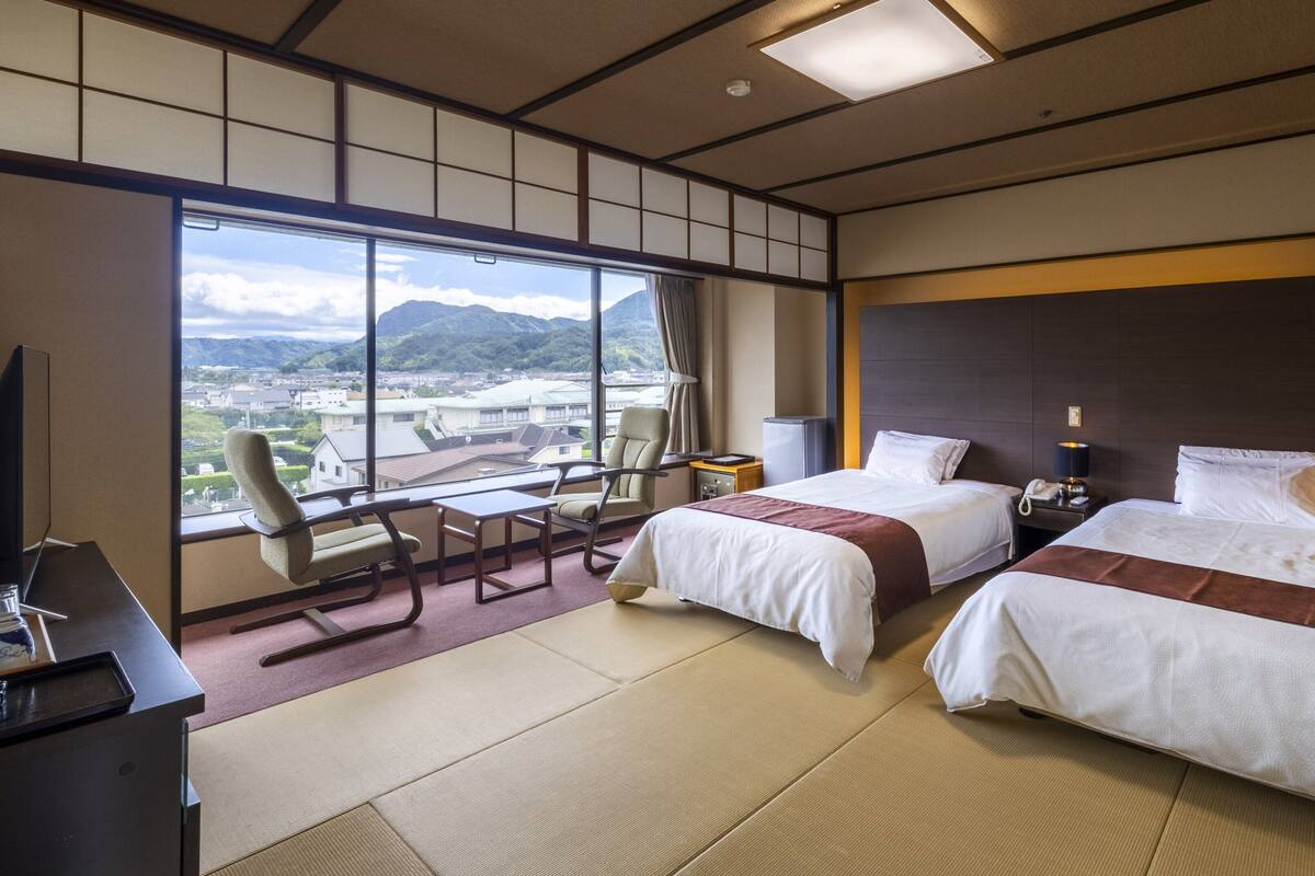 A hotel room with two beds, a desk, a chair, a TV, and a large window with a view of a city and mountains.