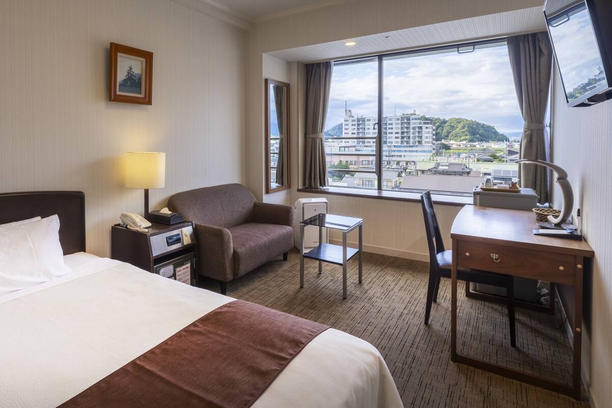 A hotel room with a bed, a desk, a chair, a lamp, a phone, a picture on the wall, and a window with a city view.