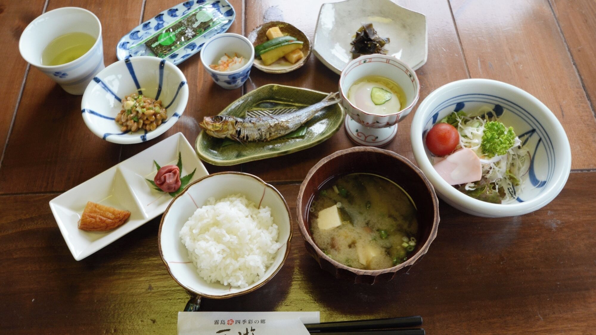A variety of Japanese dishes on a wooden table, including rice, fish, and vegetables.