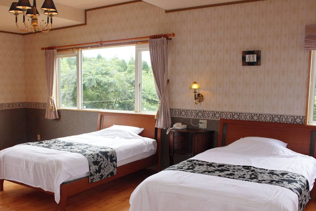 A hotel room with two beds, a wooden floor, a chandelier, a nightstand with a lamp, and a window with curtains.