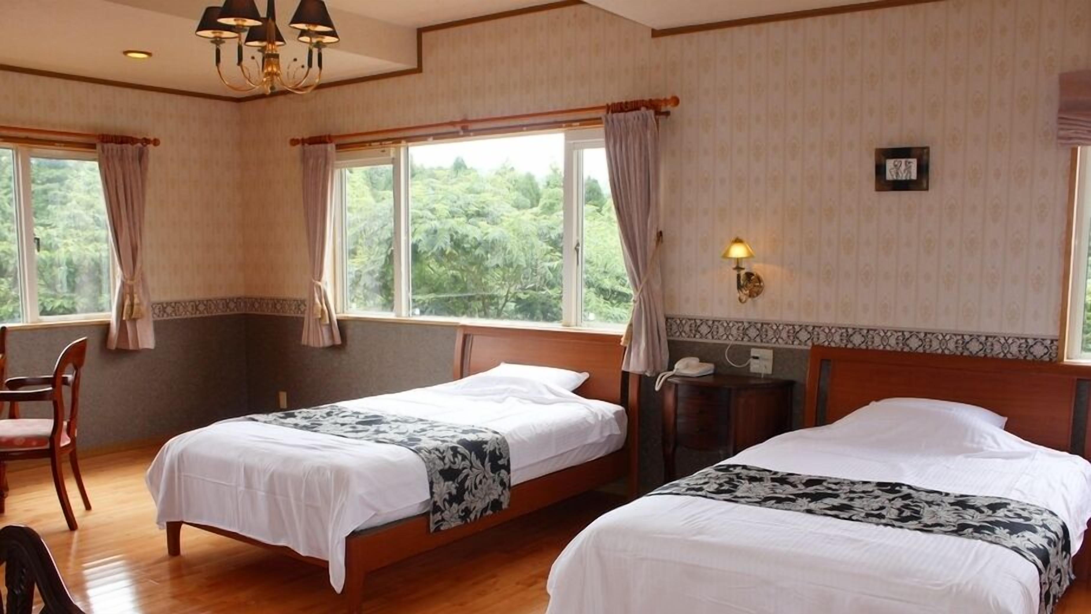 A hotel room with two single beds, a wooden floor, a chandelier, a desk with a lamp, and large windows with curtains.