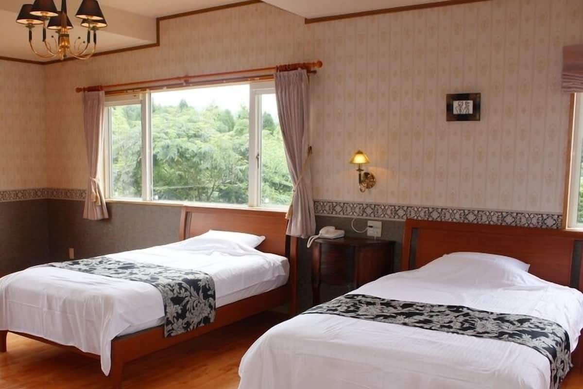A hotel room with two single beds, a wooden floor, a chandelier, a desk with a lamp, and large windows with curtains.